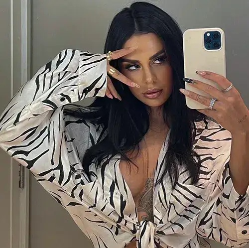 A sexy, dark haired woman poses for a selfie. She looks through parted fingers into the camera while her blouse is fully unbuttoned, revealing she is not wearing a bra.