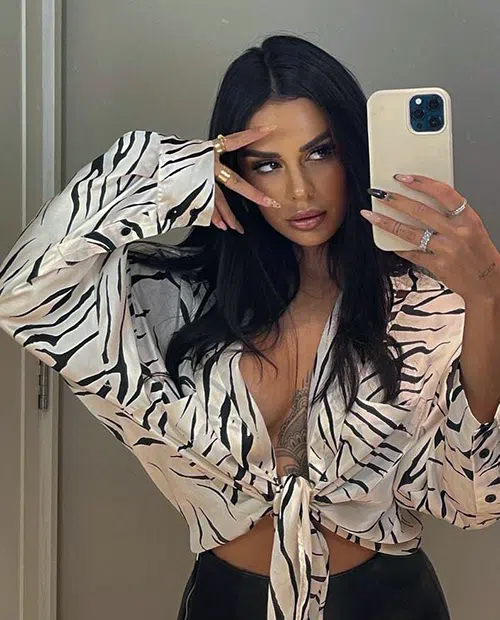 A sexy, dark haired woman poses for a selfie. She looks through parted fingers into the camera while her blouse is fully unbuttoned, revealing she is not wearing a bra.